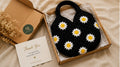 Gift box having Handmade black crochet shoulder bag featuring white and yellow daisy granny square floral pattern for everyday use.