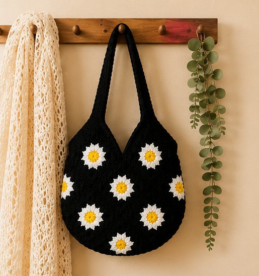 Handmade black crochet shoulder bag featuring white and yellow daisy granny square floral pattern for everyday use, hung on a wall hanger
